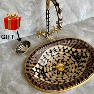 May include: A black and white patterned oval sink with a gold rim. The sink is made of a mosaic of different materials. A gold faucet with a black and white handle is attached to the sink.