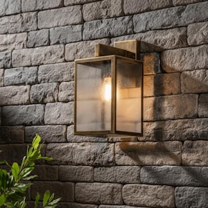 Könnte beinhalten: Eine rechteckige Außenwandleuchte mit goldfarbenem Rahmen und klaren Glasscheiben. Die Leuchte ist an einer strukturierten Backsteinmauer angebracht. Eine leuchtende Glühbirne ist im Inneren der Leuchte sichtbar und wirft einen warmen Schein. Grünes Laub ist unten links zu sehen.