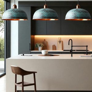 May include: Three pendant lights with teal-colored, dome-shaped shades and copper-colored interiors hang above a kitchen island. The kitchen features a wooden bar stool, a white countertop, and dark cabinets.