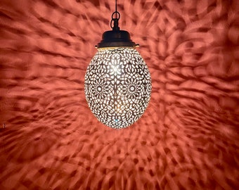 May include: A decorative hanging light fixture with a white metal shade featuring a delicate, intricate pattern. The light is on and casting a warm glow on the red wall.