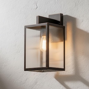 May include: A dark metal outdoor wall light with a rectangular glass enclosure. The light fitting features a visible, warm-toned bulb. It's mounted on a textured white wall, with a shadow cast to the side. The dimensions are approximately 20 cm x 15 cm.