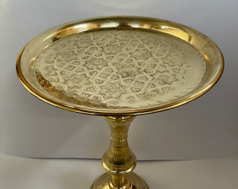 May include: A gold metal serving tray with an intricate geometric pattern on the top. The tray is attached to a tall, round pedestal with a decorative base.