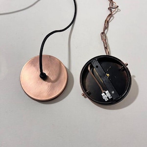 May include: Two round copper-colored ceiling light fixture canopy covers. One cover is open, revealing the electrical wiring and a black metal plate. The other cover is closed.