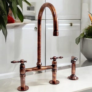 Puede incluir: Un grifo de cocina de cobre con un diseño de puente y un rociador lateral. El grifo tiene dos manijas y un caño de arco alto.