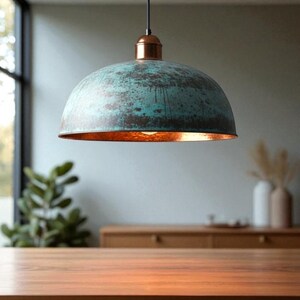 May include: A dome-shaped pendant light with a weathered teal and copper finish. The interior of the shade has a warm, orange glow. The light hangs from a black cord and copper fixture, suitable for a kitchen or dining room.