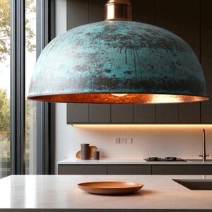 May include: A large, dome-shaped pendant light fixture with a weathered teal and copper finish. The interior of the shade has a warm, orange glow. The fixture hangs above a kitchen countertop with a wooden plate.