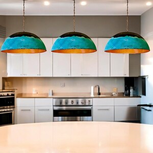May include: Three turquoise and copper pendant lights hang over a white kitchen island. The lights have a dome shape and are suspended from a chain.