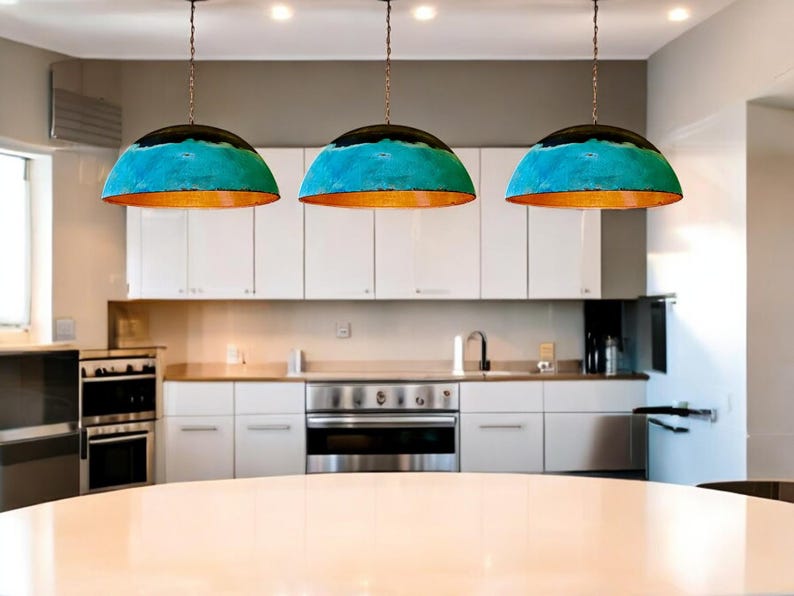 May include: Three turquoise and copper pendant lights hang over a white kitchen island. The lights have a dome shape and are suspended from a chain.
