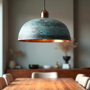 May include: A large, dome-shaped pendant light with a weathered teal and copper finish. The interior of the shade has a warm, orange glow. The light hangs above a wooden dining table, suggesting a dining room setting.
