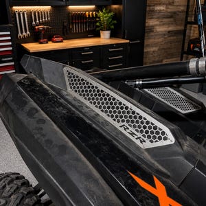 以下が含まれることがあります： オレンジ色のアクセントが付いた、黒とシルバーのRZRサイドバイサイド車。ハニカムグリルデザインとシルバーの「RZR」の文字が特徴です。画像はガレージで撮影されました。