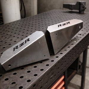 Puede incluir: Dos componentes metálicos plateados con la palabra "RZR" impresa en negro. Las piezas están anguladas y perforadas con agujeros. Se encuentran sobre una mesa metálica con un patrón de rejilla. Un tanque de gas verde es visible en el fondo.