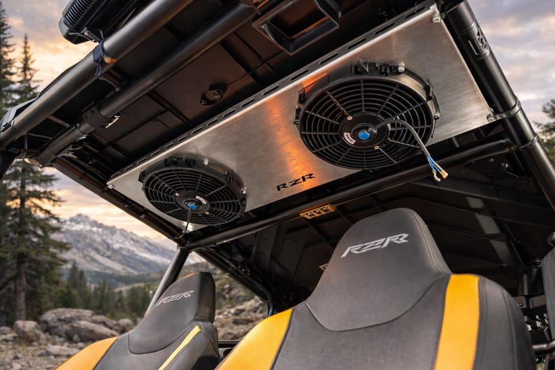 May include: An overhead view of a RZR off-road vehicle interior. Two black cooling fans are mounted on a silver panel, with the RZR logo visible. The seats are black with yellow accents. The background shows a mountain landscape.