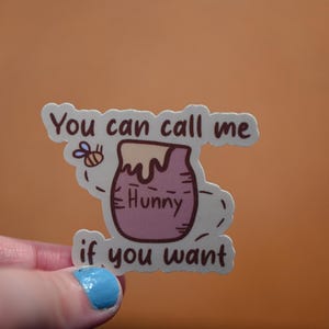 May include: A sticker featuring a cartoon honey pot with the word "Hunny" and the phrase "You can call me if you want." A small bee is also included in the design. The sticker is held by a hand with blue nail polish.