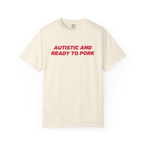 May include: Cream-colored t-shirt with the red text "AUTISTIC AND READY TO PORK" in a bold, sans-serif font. The shirt has a classic crew neck and short sleeves. The fabric appears to be a soft, comfortable material.