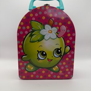 May include: A pink metal lunchbox with a teal handle and a cartoon apple design. The apple has a smiling face, a white flower, and a small worm. The background is covered in colorful polka dots.