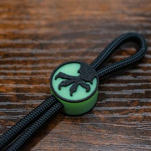 May include: A black and green cord with a round, green bead featuring a black, stylized, tentacled creature design.