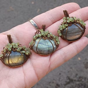 May include: Three handmade gemstone pendants. Each pendant features a polished, oval-shaped labradorite stone wrapped in copper wire. The wirework is adorned with small, green peridot crystals. The pendants are displayed on a hand.