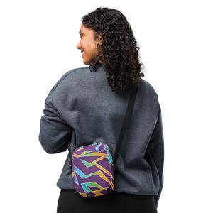Peut inclure: Un sac bandoulière violet avec un motif géométrique aux couleurs jaune, vert et bleu. Le sac est porté sur l'épaule avec une sangle noire. La personne sur l'image porte un sweat-shirt gris.