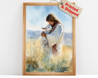 May include: A framed watercolor print depicts a person in white robes embracing a lamb in a field of tall grass. The background features rolling hills and a blue sky. The frame is made of light brown wood.