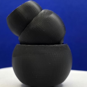 May include: Three black, spherical, 3D-printed objects stacked on top of each other. The bottom object is a complete sphere, while the top two are open at the top. The objects are on a white surface with a blue background.