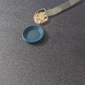 May include: A teal-colored, circular container and a gray tool with a small amount of a brown substance. The objects are on a dark gray, textured surface. The word "ANKER" is visible in the background.