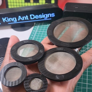 May include: A collection of black circular filters with mesh centers, varying in size, are displayed. The filters are held in a hand, with a cutting mat and a pixelated display in the background. The text "King Ant Designs" is visible.