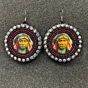 May include: A pair of round earrings featuring a Native American portrait. The earrings have a black beaded border with red and clear crystal accents. The central image is a colorful portrait of a Native American man wearing a headdress.
