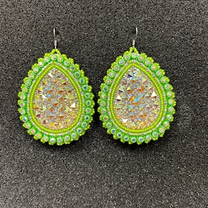 May include: A pair of handmade, teardrop-shaped earrings. The earrings feature a vibrant neon green beaded border and a sparkling, iridescent center. The earrings are designed with a hook closure.
