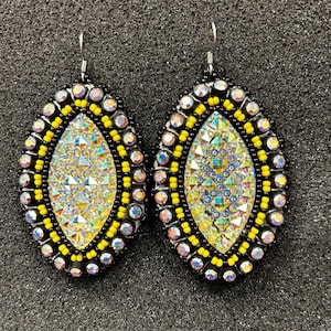May include: A pair of teardrop-shaped earrings. The earrings feature a central iridescent design surrounded by yellow, black, and clear beads. The earrings have silver-colored hooks.