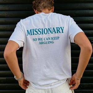 May include: White t-shirt with the word "MISSIONARY" printed in teal, along with the phrase "SO WE CAN KEEP ARGUING." The shirt is short-sleeved and appears to be made of cotton. The person is wearing white pants and a silver bracelet.