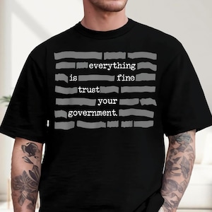 May include: Black t-shirt with the text "everything is fine trust your government" in a typewriter font. The text is arranged in a block format with gray horizontal lines above and below the text.