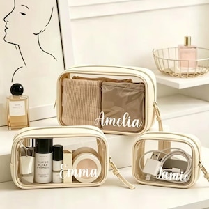 Puede incluir: Tres neceseres transparentes con ribetes y cremalleras color crema. Las bolsas están etiquetadas con "Amelia", "Emma" y "Jamie" en escritura blanca. Las bolsas contienen maquillaje, artículos de tocador y accesorios. Las bolsas están sobre una superficie blanca.