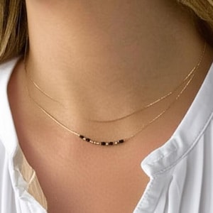 May include: A delicate gold necklace with black beads. The necklace is worn against a white shirt. The image shows a close-up of the neck and upper chest.