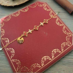 May include: A gold chain bracelet with a red stone charm and a gold charm with the letter E. The bracelet is on a red velvet box with gold trim.