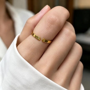 Pode incluir: Um anel aberto em tom dourado com duas extremidades planas, cada uma gravada com uma letra individual. Uma extremidade exibe a letra F e a outra extremidade exibe a letra S. O anel tem um acabamento liso e polido com um design simples.