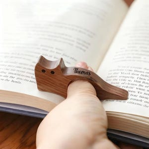 Può includere: Un fermapagina in legno a forma di gatto, con il nome "Thomas" inciso. Il supporto marrone ha due piccoli fori rotondi per gli occhi. È progettato per essere infilato su un dito per tenere aperto un libro.