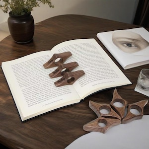 Può includere: Segnalibri a pollice in legno con i nomi "Amy", "Amelia" e "Sophia" incisi, appoggiati su un libro aperto. Sono presenti anche segnalibri in legno a forma di stella. Gli oggetti sono su un tavolo di legno scuro.