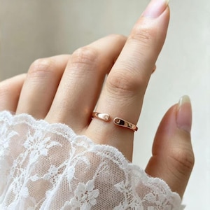 Puede incluir: Un anillo abierto de oro rosa con una banda lisa y pulida que presenta una pequeña abertura. Los extremos del anillo están aplanados y grabados con las letras mayúsculas A y J, una a cada lado de la abertura, creando una pieza de joyería personalizada.