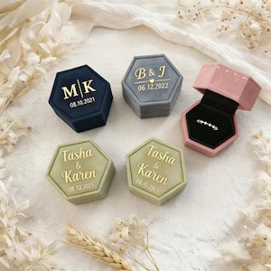 Könnte beinhalten: Fünf sechseckige Ringboxen in verschiedenen Farben: Marineblau, Grau, Rosa und zwei Grüntönen. Jede Box ist mit goldenen Buchstaben personalisiert, einschließlich Initialen, Namen und Daten. Eine rosa Box ist geöffnet und zeigt einen Ring.