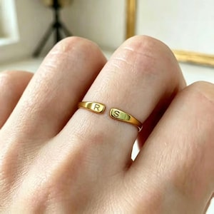 May include: A gold-toned open ring featuring two flat, rounded ends. Each end is engraved with a single capital letter, with the letter R on the left side and the letter S on the right side. The band has a smooth, polished finish throughout.