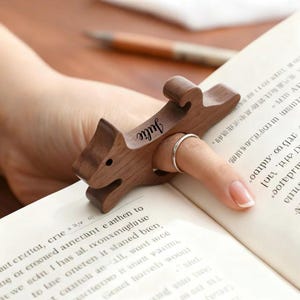 Può includere: Un fermalibri in legno a forma di lupo, con il nome "Julie" inciso. Il supporto è progettato per essere tenuto su un dito durante la lettura. Il libro aperto mostra il testo sulle sue pagine.