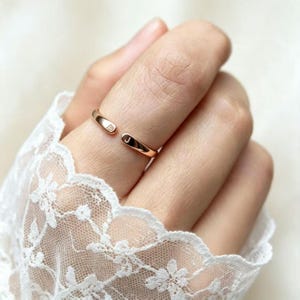 Puede incluir: Un anillo abierto de oro rosa con una banda lisa y pulida, que presenta una pequeña letra A grabada en un extremo y una letra J grabada en el otro. El anillo se lleva puesto en un dedo, destacando su diseño sencillo y acabado metálico.