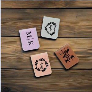 Könnte beinhalten: Vier personalisierte Leder-Lesezeichen mit verschiedenen Designs. Ein Lesezeichen ist rosa mit den Initialen "M|K" in Schwarz. Ein weiteres Lesezeichen ist hellbraun mit einem Kranz-Design und dem Namen "Amy" in Schwarz. Ein drittes Lesezeichen ist weiß mit einem Kranz-Design und dem Namen "Sophia" in Schwarz. Das letzte Lesezeichen ist braun mit dem Namen "Martin" in Schwarz und den Initialen "M|T" in Schwarz.