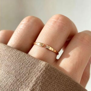 May include: A rose gold-toned open ring featuring a polished finish. One end of the band is engraved with the capital letter L, while the opposite end displays a small, detailed paw print icon. The ring has a slim, minimalist band design.