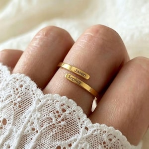 May include: A gold-coloured bypass ring featuring two open ends. Each end of the ring is engraved with a name, with the top end displaying the name Jason and the bottom end displaying the name Sophia in a clean, sans-serif font style.