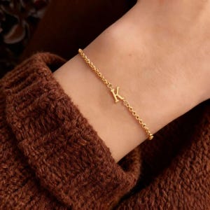 Puede incluir: Una pulsera dorada con una cadena delicada y un dije con la letra "K". La pulsera se lleva en la muñeca, con un suéter de punto marrón en el fondo. Una joya elegante.