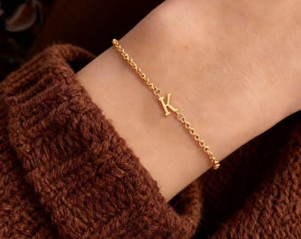May include: A gold bracelet featuring a delicate chain and a letter "K" charm. The bracelet is worn on a wrist, with a brown knitted sweater in the background. The jewelry is a stylish accessory.
