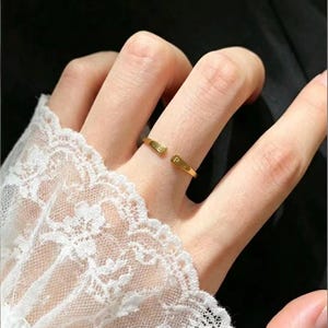 May include: A gold-toned open ring featuring two flat ends, each engraved with a single capital letter. One end displays the letter E and the other end displays the letter P. The ring has a smooth, polished finish and a simple, adjustable band design.