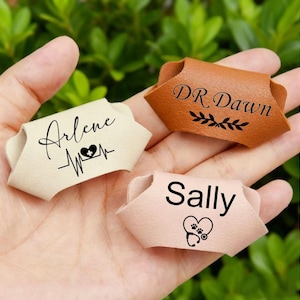 May include: Three personalised leather name tags in tan, brown, and pink. The tags feature names and medical-themed graphics, including a heartbeat, a heart with a cross, and a heart with paw prints and a stethoscope. The tags are held in a hand.