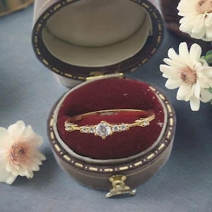 Op de afbeelding: Een gouden ring met een enkele grote, heldere steen en kleinere stenen aan beide kanten, geplaatst in een ringdoos met een rood fluwelen interieur.